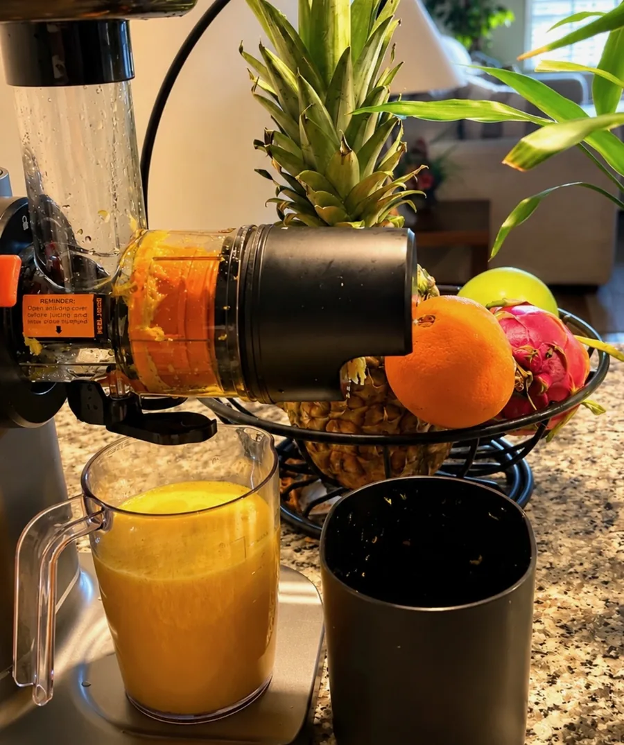 A cold-press juicer pouring fresh orange juice into a glass pitcher, with pineapple, oranges and dragonfruit in a bowl behind it
